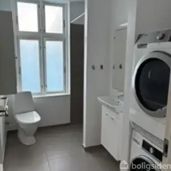 Vaskemaskine og tørretumbler er stablet og placeret ved siden af en lille vask. Toilet er placeret nær et vindue, og rummet har hvide vægge.