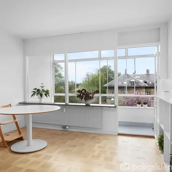 Et moderne rum med et rundt spisebord og træstol i forgrunden. To planter står på radiator under store vinduer, der giver udsigt til en boligbygning og grønne træer.