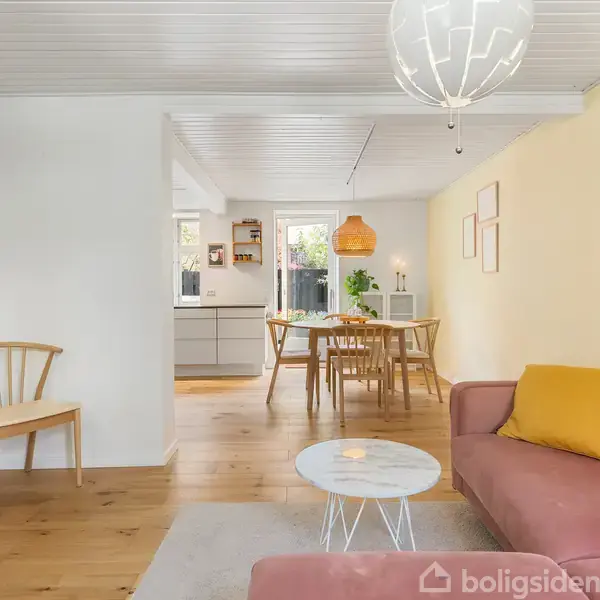 Et hvidt køkken med moderne apparater; en espressomaskine står ved siden af en indbygget ovn. Køkkenets vægge har hvide fliser. Runde stole og hylder med glas i baggrunden.