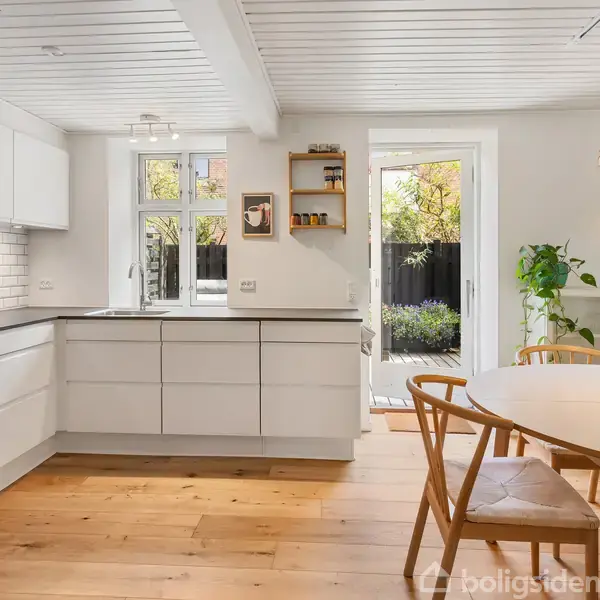 Køkken med hvide skabe og sort bordplade, står uden aktivitet, omgivet af trægulv og spiseplads. Vindue og terrassedør åbner til en have. Moderne indretning med køkkenlampe og planter.