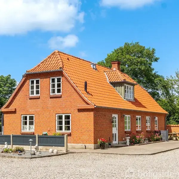 En rød murstensvilla med orange tegltag står i en solrig have. Huset har flere vinduer og en indgangsdør med blomsterdekorationer ved siden af en grusbelagt gårdsplads.
