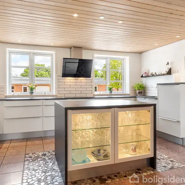 Et moderne køkken med en glasvitrineskab, der viser glas og tallerkner. Skabe omgiver rummet med en vægmonteret TV-skærm. Store vinduer giver udsigt til en grøn have.