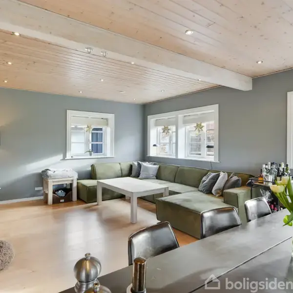 Stor L-formet grøn sofa står i et stuehjørne, flankeret af to vinduer med udsigt til haven. Rummets atmosfære er lys med et træloft og diskret dekoration.