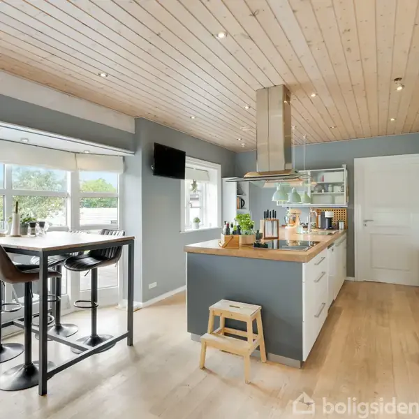 Et moderne køkken med ø-bar, barstole og køkkenø, udstyret med komfur og emhætte. Vinduer giver udsigt til en have. Lyst træloft og gulve giver rummet en varm atmosfære.