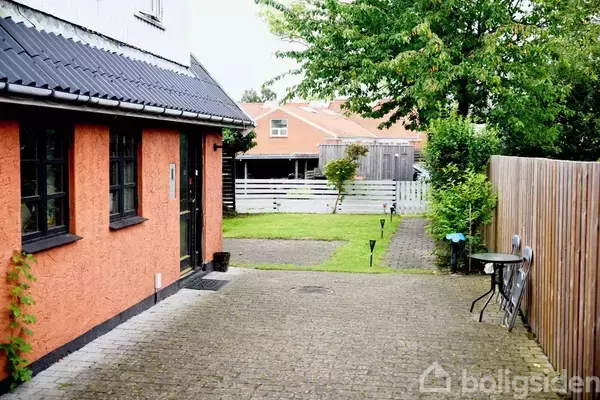 Et hus med orange facade og sorte vinduer står ved siden af en brolagt gårdsplads. En have med græs og hegn ses i baggrunden ved siden af et bord og to stole.