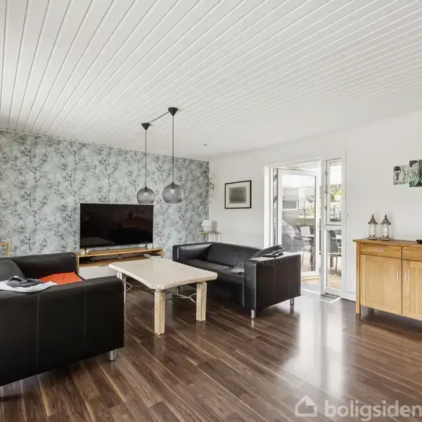 Stue med sorte lædersofaer omkring et træbord, fjernsyn på blomstret væg, to loftslamper. Trægulv, hvide vægge og døre mod terrasse. Skænk med dekorationer. Text: "Home Sweet Home".