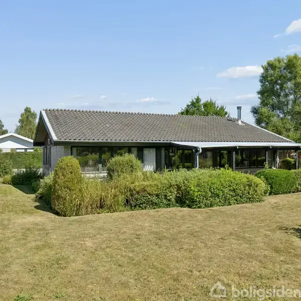 Et én-etagers hus med fladt tag står stille omgivet af en velplejet have med græsplæne og buske, under en klar, blå himmel med få skyer.