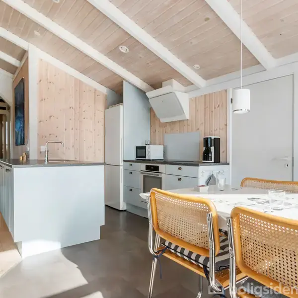 Et lyst køkken med spisebord og stole, køkkenø og moderne apparater. Trævægge og loftsbjælker skaber en hyggelig atmosfære i et veloplyst rum med store vinduer.