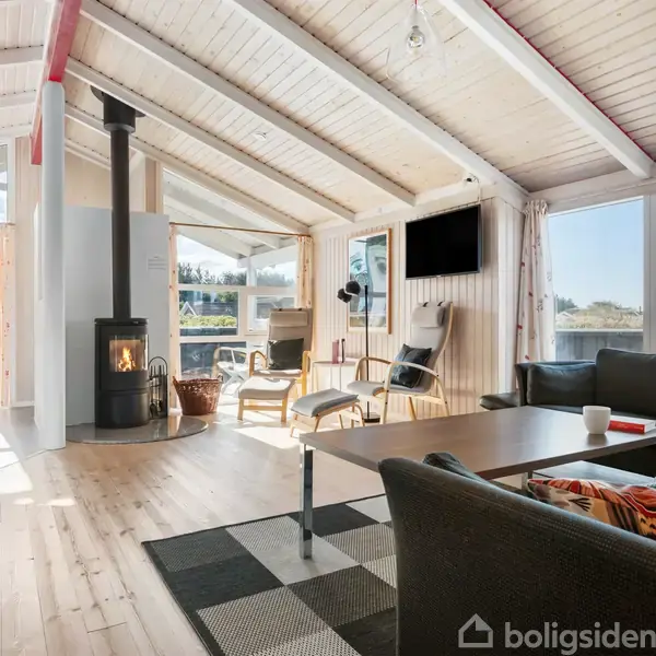Brændeovn opvarmer en lys stue med trægulve og loft til kip, omgivet af store vinduer. Sofa, bord og lænestole skaber en hyggelig atmosfære. TV hænger på væggen.