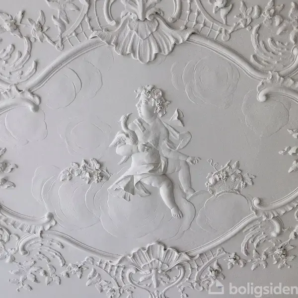 En detaljeret stukdekoration pryder loftet, hvor en engel sidder omgivet af blomsterranker. Dette kunstværk er lavet i hvidt relief, hvilket skaber en elegant, barok atmosfære.