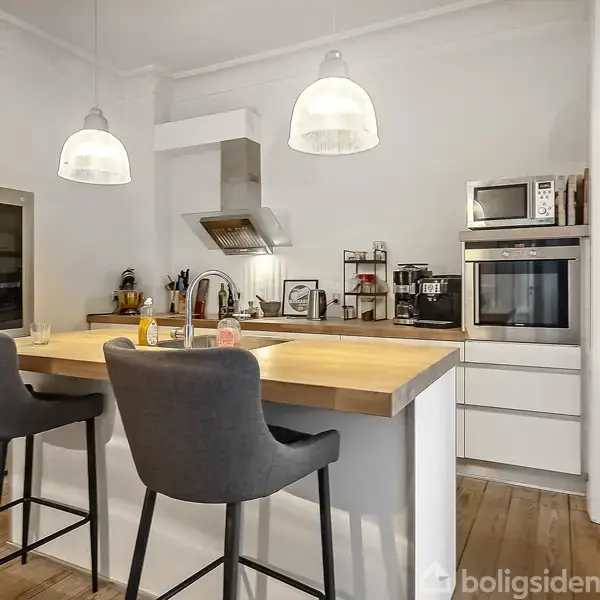 Køkken med to barstole ved en ø, omgivet af køkkenfaciliteter som indbygget ovn, mikrobølgeovn, komfur og køleskab, under loftslamper i en moderne indretning.