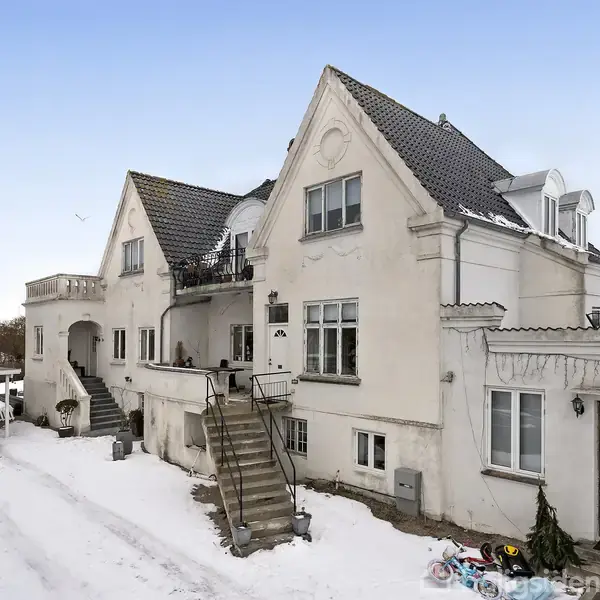 Hvid villa med karakteristisk gavl, trapper foran indgang, omgivet af snedækket grund. I baggrunden ses en carport og flere store træer på en klar dag.