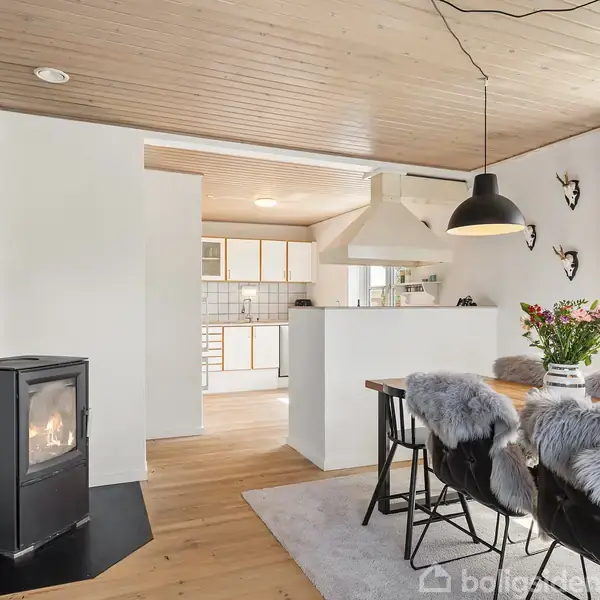 En brændeovn står tændt i en hyggelig stue, hvor et spisebord med stole dækket af pels ligger i forgrunden. Køkkenet ses i baggrunden med lyse træpaneler.