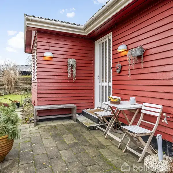 Rød træhusfacade med to udendørs væglamper. En lille terrasse har bord og stole. Blomsterkrukke står ved siden af. Græsplæne og hegn i baggrunden.