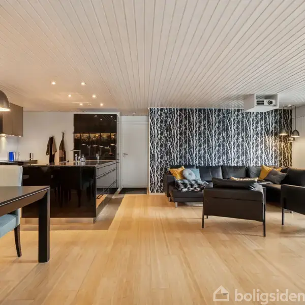 Stort åbent rum med træparketgulv. En sort sofa står ved en væg med skovmotiv-tapet. Et spisebord med blomster og lamper hænger over køkkenområdet i baggrunden.