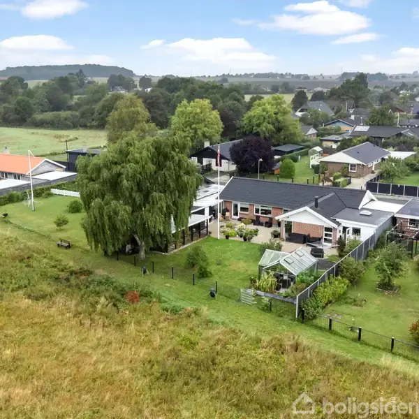Huse ligger langs en række i et grønt, landligt område. Lofter er sorte og røde, med velplejede haver. Omkringliggende marker og træer er synlige, skaber en fredelig atmosfære.