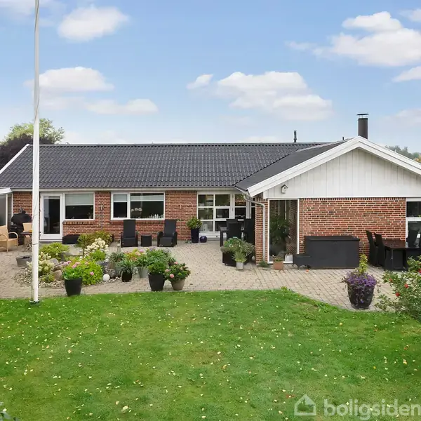 Rødt murstenshus med sort tag står frit med flisebelagt terrasse. Flere blomsterpotter og stole ses foran huset. Græsplæne med træer omkranser området under en skyfri, blå himmel.