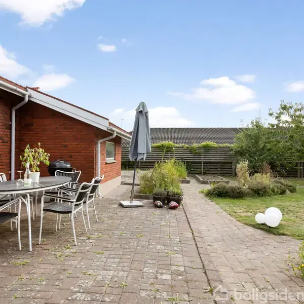 Haven indeholder et terrasseområde med et bord og stole ved siden af husets murstensvæg. Der står en parasol, og græsplænen har spredte planter samt en hæk i baggrunden.
