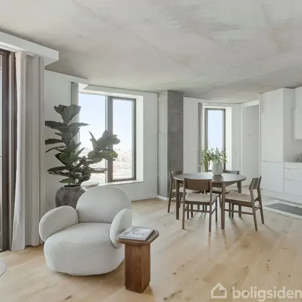 Stue med en designerstol ved siden af et lille bord og en stor plante. Bagved ses spisebord med stole og køkkenområde i lys, moderne stil med store vinduer.