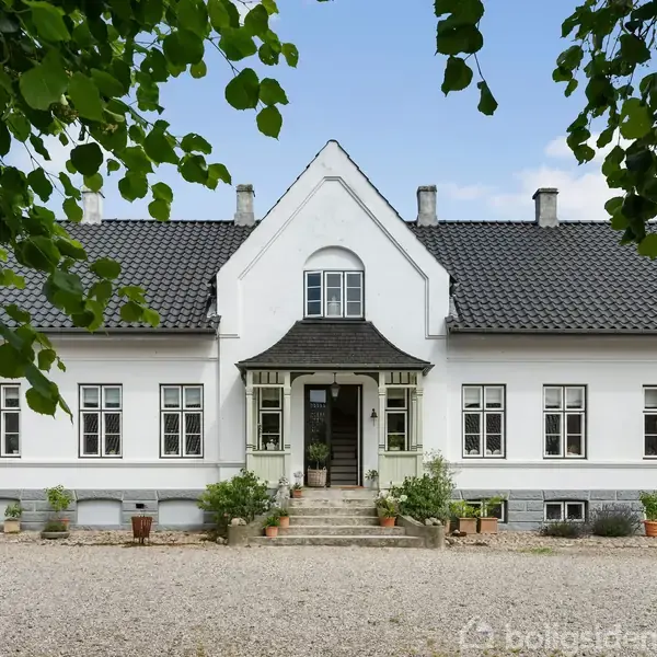 Elegant hvid villa med sort tegltag, står stille i en gårdsplads omkranset af grus og grønne planter, skaber en fredfyldt, landlig atmosfære.