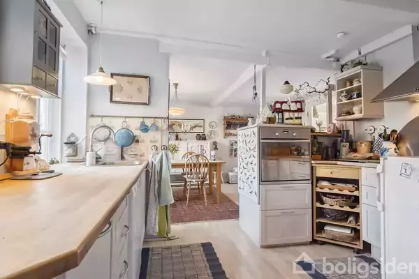 Et lyst køkken med træbordplade og hvide skabe overrinder i rummet. Spisebord med stole kan ses i baggrunden. Hylderne er fyldt med køkkenartikler og dekorationer.