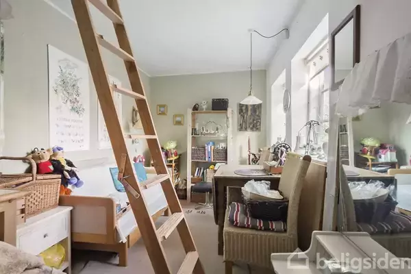 En stige står op ad en væg i et rum med beige vægge og en skrivebordspult fyldt med genstande. Der er også en sofa med bamser og reoler i baggrunden.