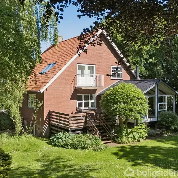 Et lyst og rummeligt orangeri med store glaspartier, der indrammer en grøn have. En blød sækkestol og et bord med blomster står på trægulvet.