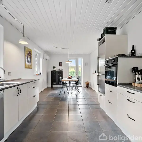 Lyst køkken med hvide skabe og stål-apparater. Et rundt bord med stole ses i baggrunden nær vinduer. Flisegulv og loftslampe i moderne stil.