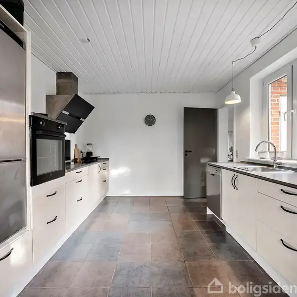 Et moderne køkken med hvide skabe og en indbygget ovn i aktion. Rummet har fliser på gulvet, en stålvask under et stort vindue, og enkel belysning.