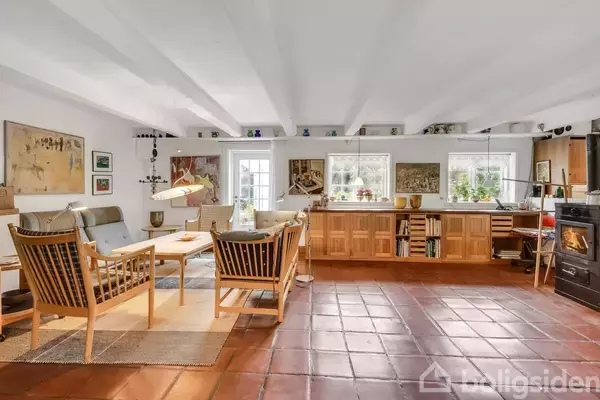 Stue med træmøbler hviler på et tæppe, omgivet af kunstværker på væggene. Store vinduer giver lys, og brændeovn står til højre. Flisegulv og synlige bjælker præger rummet.