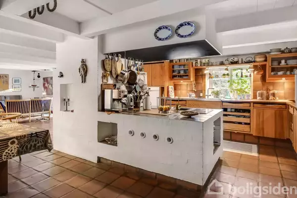 Et køkken med hvide murstensvægge og træskabe. Gryder og pander hænger over køkkenøen. I baggrunden ses en spisestue med træmøbler. Vinduer giver naturligt lys.