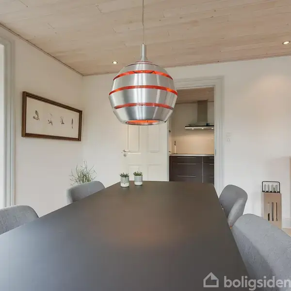 Pendellampe hænger over sort spisebord. Stuen har grå stole, en hvid dør fører til køkkenet, og en brændeovn står til højre. En stor plante samt et maleri pynter væggen.
