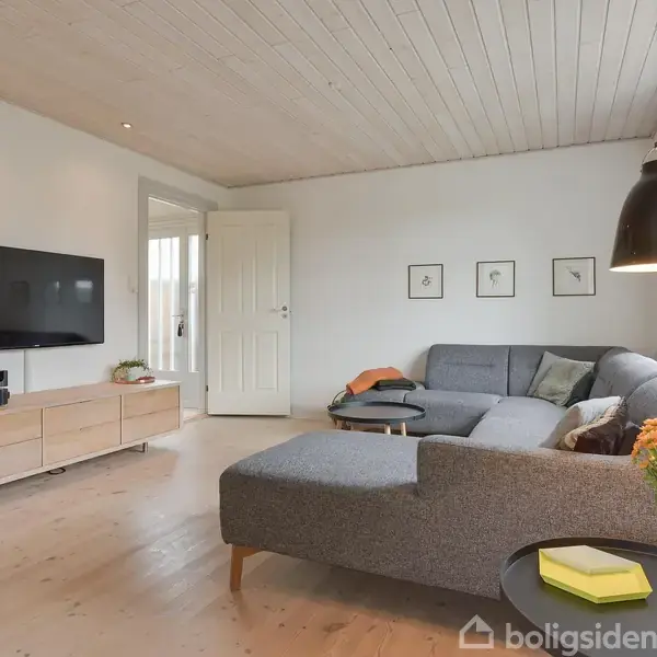 En grå hjørnesofa står i en lys stue ved siden af et sofabord med blomster. TV og skænk ses på venstre væg, træloft over.
