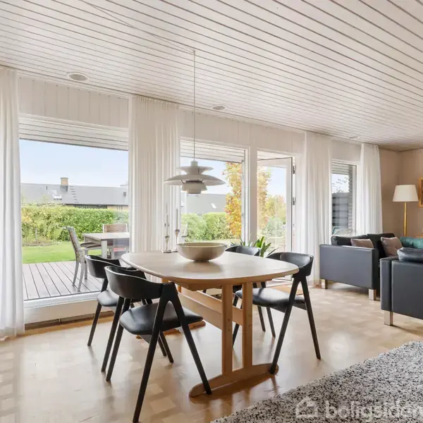 Et moderne spisebord med fire stole står i en lys stue. Glasdøre åbner ud mod en terrasse og have. Et tæppe og sofaer kompletterer rummet.