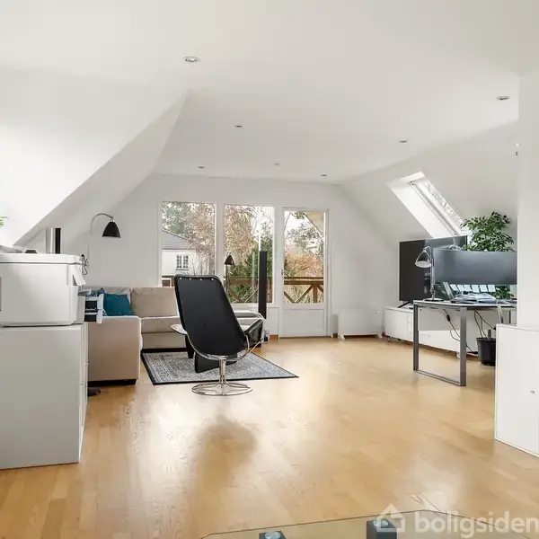 Stue med sofa og lænestol placeret på tæppe; lysstrømmende gennem store vinduer. Maleri hænger på væggen. Stuen er rummelig og har trægulv.