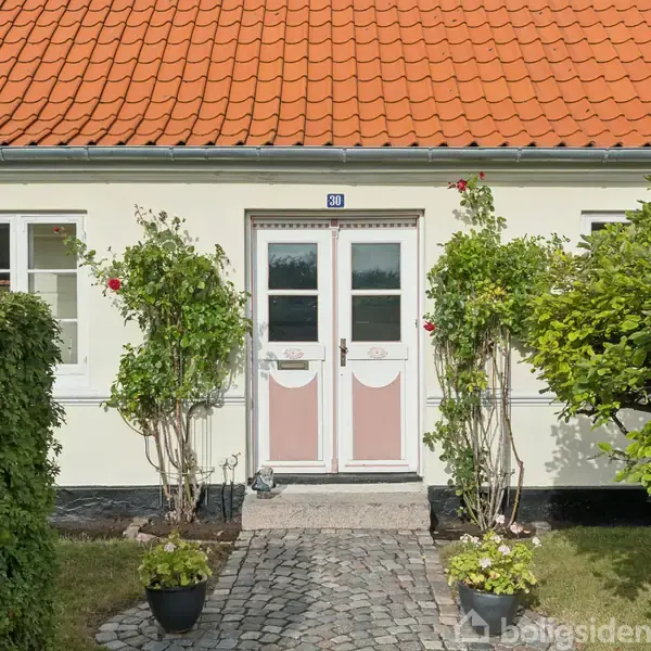 Dør omkranset af blomstrende planter står centralt i en hvid murstensfacade med rødt tegltag. Flisebelagt sti og grønne buske danner forhave, mens et rundt blåt husnummer ses over døren.