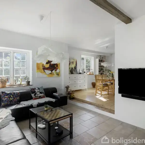 Sort hjørnesofa står i en lys stue med hvidmalede vægge. Et tv hænger på væggen, mens spisebord og stole står i baggrunden. Indretningen inkluderer farverigt kunstværk.