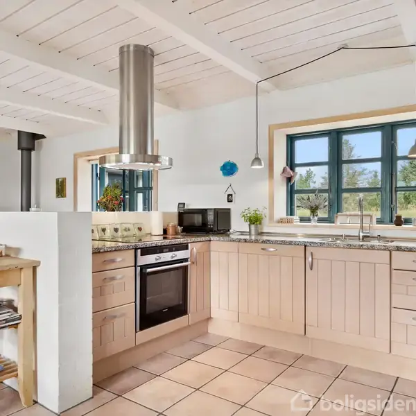 Et moderne køkken med træskabe og granitbordplader. En ovn og mikroovn står ved siden af en udtrækssugemekanisme. Et vindue giver udsigt til naturen udenfor.