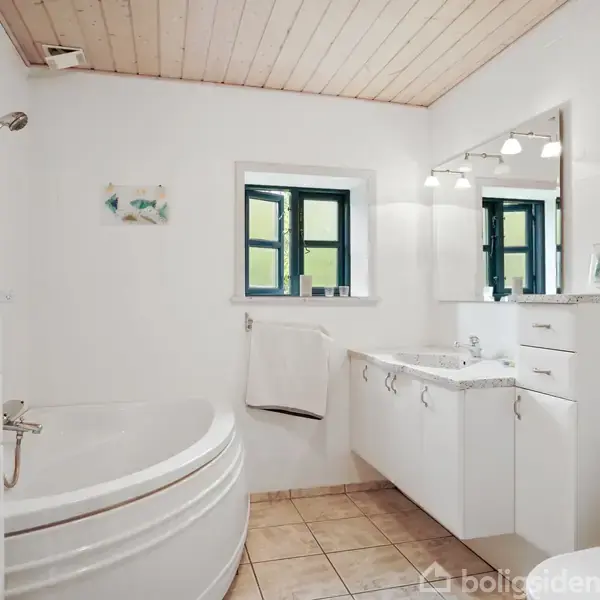 Badeværelse med hvidt badekar i venstre hjørne, hvide skabe og vask med spejl på højre side. Vindue med sorte rammer i midten. Flisegulv og træbeklædt loft.