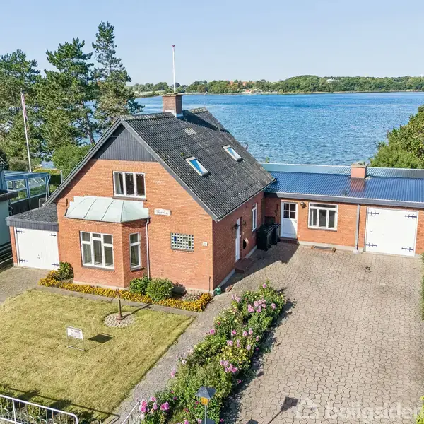 Murstenshus med sort tag står ved vandkanten, omgivet af en have med blomster og en stenbelagt indkørsel. I baggrunden ses en stor sø og træer.