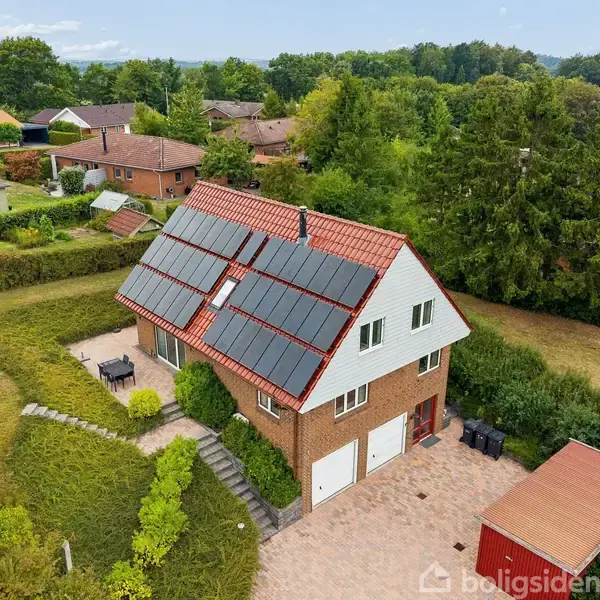 Et murstenshus med solpaneler på taget, omgivet af en grøn have med en terrasse og en rød garage. Det ligger i et beboelsesområde med flere huse og træer omkring.