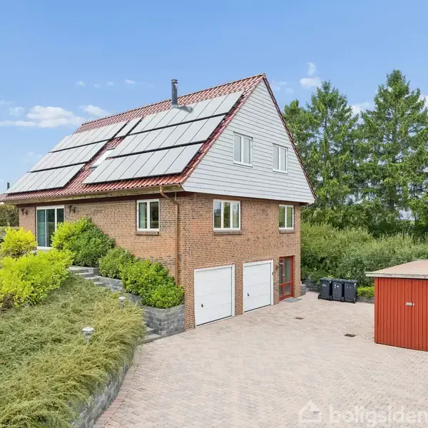 Et to-etagers hus har solpaneler på taget og to garageporte. Det er omgivet af grønne buske og træer, med en rød udvendig bygning til højre på en flisebelagt indkørsel.