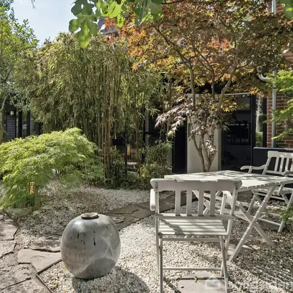 To hvide metalstole står ved et havebord på en terrasse omgivet af frodig vegetation og stenbelægning. En stor rund vase er placeret i midten af grusstien.
