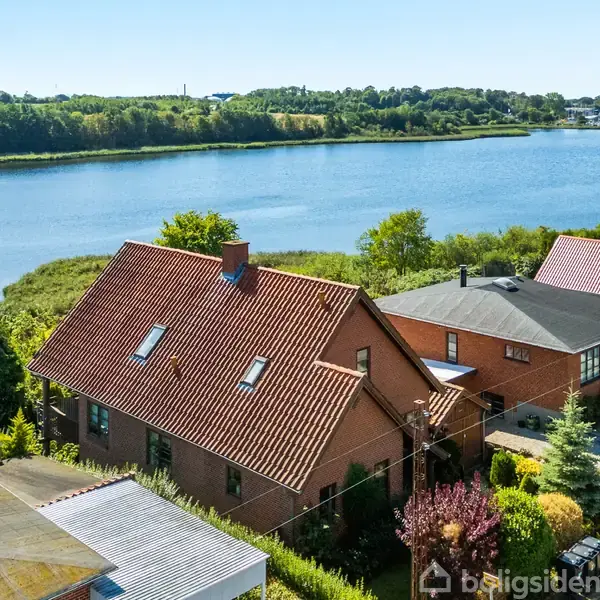 Murstenshus med rødt tegltag står ved en sø, omgivet af frodig vegetation og andre huse. I baggrunden ses en række træer på den modsatte bred.