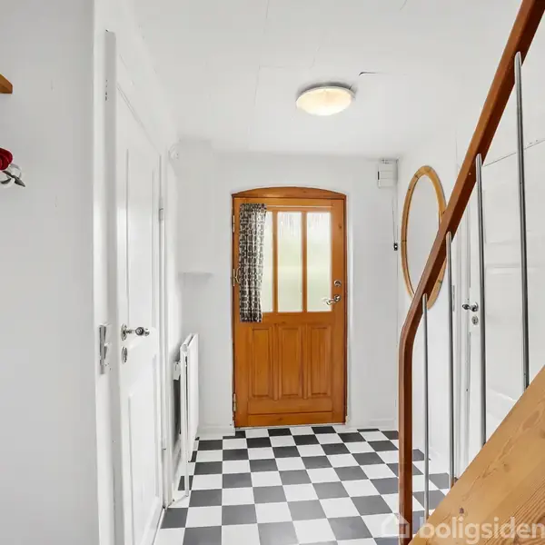 Trædør står i en entre med skakternet gulv. En trappe med trærækværk til højre, mens en hylde med bøjler hænger til venstre. Hvidt interiør omgiver rummet.