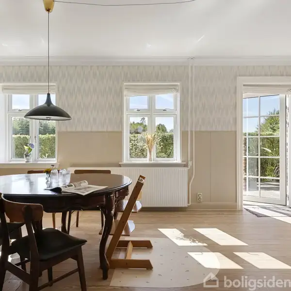 Et rundt spisebord med stole står i et lyst rum fyldt med dagslys. Store vinduer og en dør fører ud til en terrasse med udsigt til grønt område.
