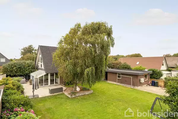 Huset har et skrånende tag og en stor glasudbygning, ligger i en grøn have med træer og buske. Til højre er der en mindre bygning.