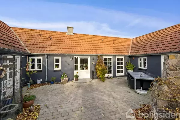 En gårdsplads med brosten omringet af sortmalede bygninger med røde tegltage. Hoveddøren er omgivet af potteplanter, og der er et bord langs væggen. Sollys skinner på området.