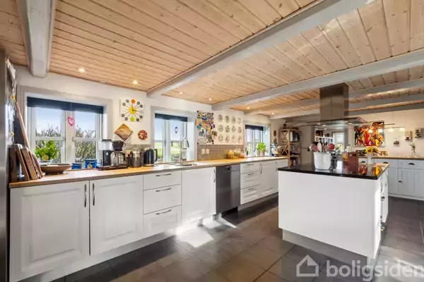 Et lyst køkken med hvide skabe og sort bordplade. Køkkenøen er centralt placeret. I baggrunden er der vinduer med udsigt, dekorationer på væggene og indbyggede lofter med spots.