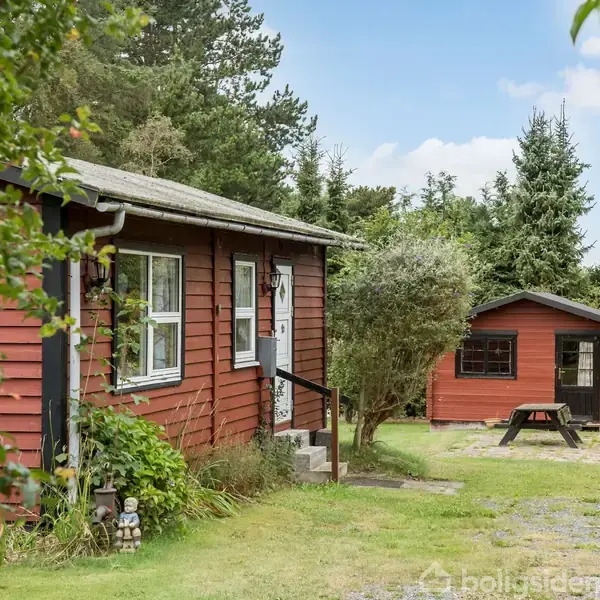 Rødt træhus med hvide vinduer står stille i en grøn, frodig have. Til højre ses et lille skur og en træbænk på en grusbelagt sti omgivet af træer.